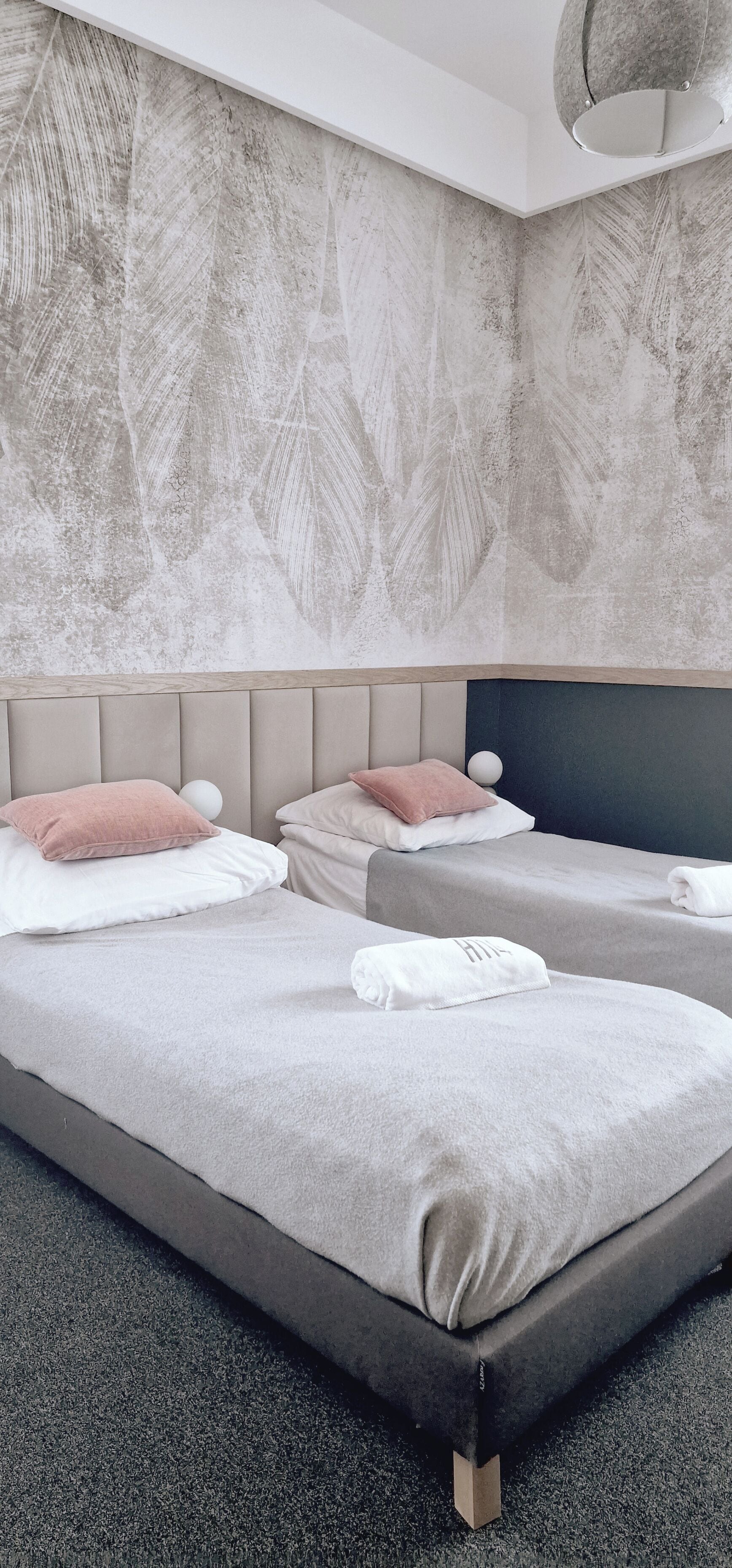 A modern hotel room with two beds, a textured leaf-patterned wall, and a ceiling light fixture.