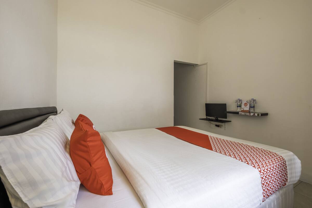 A single bed with white bedding and an orange pillow, a television on a stand, and a shelf with toiletries.