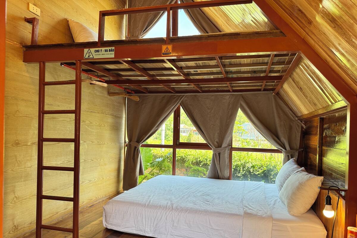 A single bedroom with a bunk bed, a wooden ladder, a window with curtains, and a bed with white bedding.