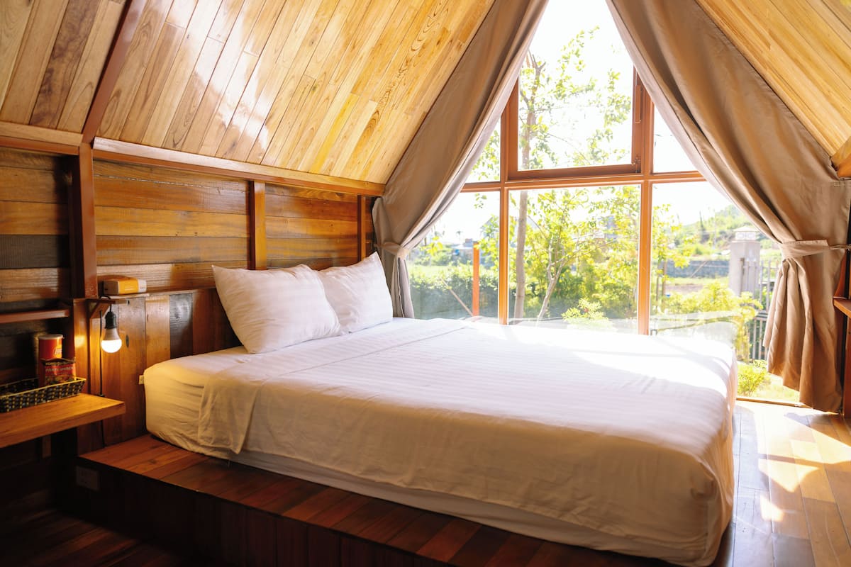 A bedroom with a wooden ceiling, a large bed with white linens, a nightstand with a lamp, and a window with curtains.