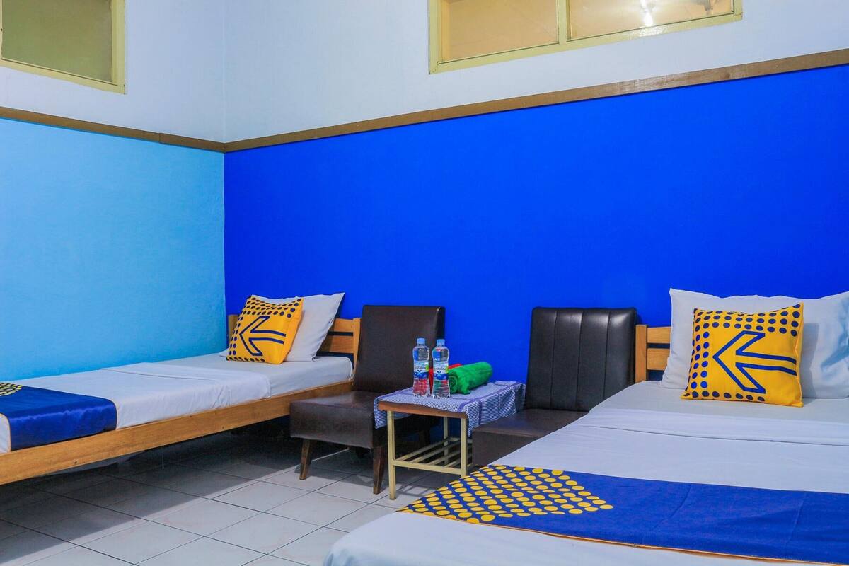 Una habitación de hotel con dos camas individuales, una mesita con una botella y una toalla verde, y una pared azul.
