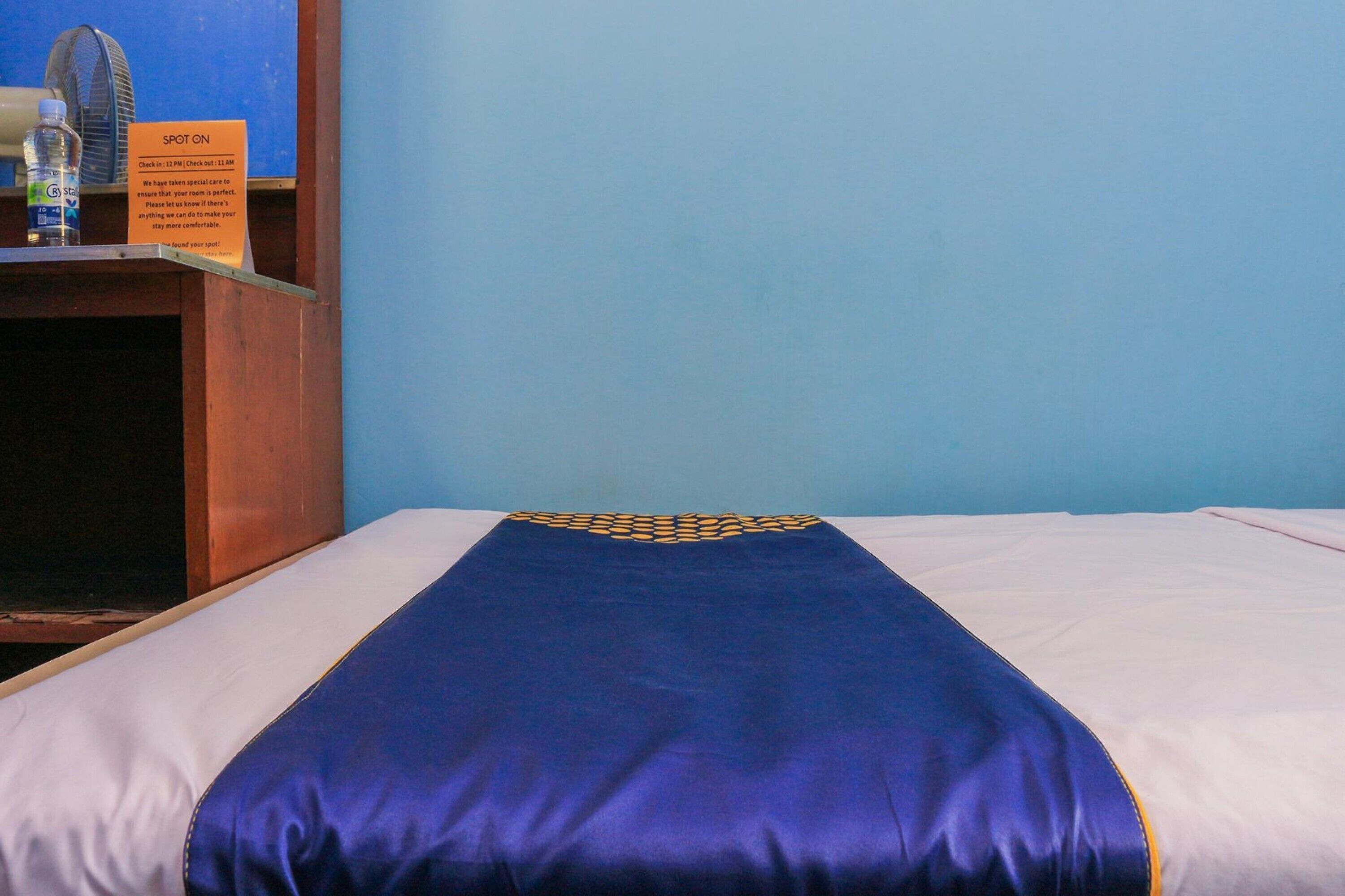 A hotel room with a bed, a wooden nightstand, a fan, and a sign indicating check-in times.