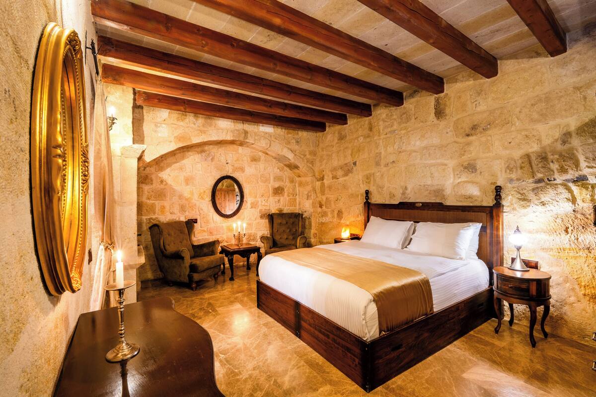 Un dormitorio con pared de piedra, vigas de madera, cama, dos sillones, una mesa con velas y un espejo.