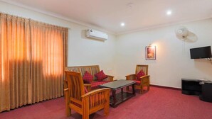 A room with a sofa, two chairs, a small table, an air conditioning unit, a wall-mounted TV, and a large window with curtains. - RamKripa Inn (Karwi)
