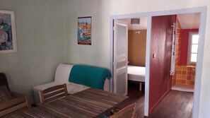 Una habitación pequeña y acogedora, con una mesa de madera, un sofá blanco y una cama. Hay un cartel en la pared y una puerta que da a otra habitación.
