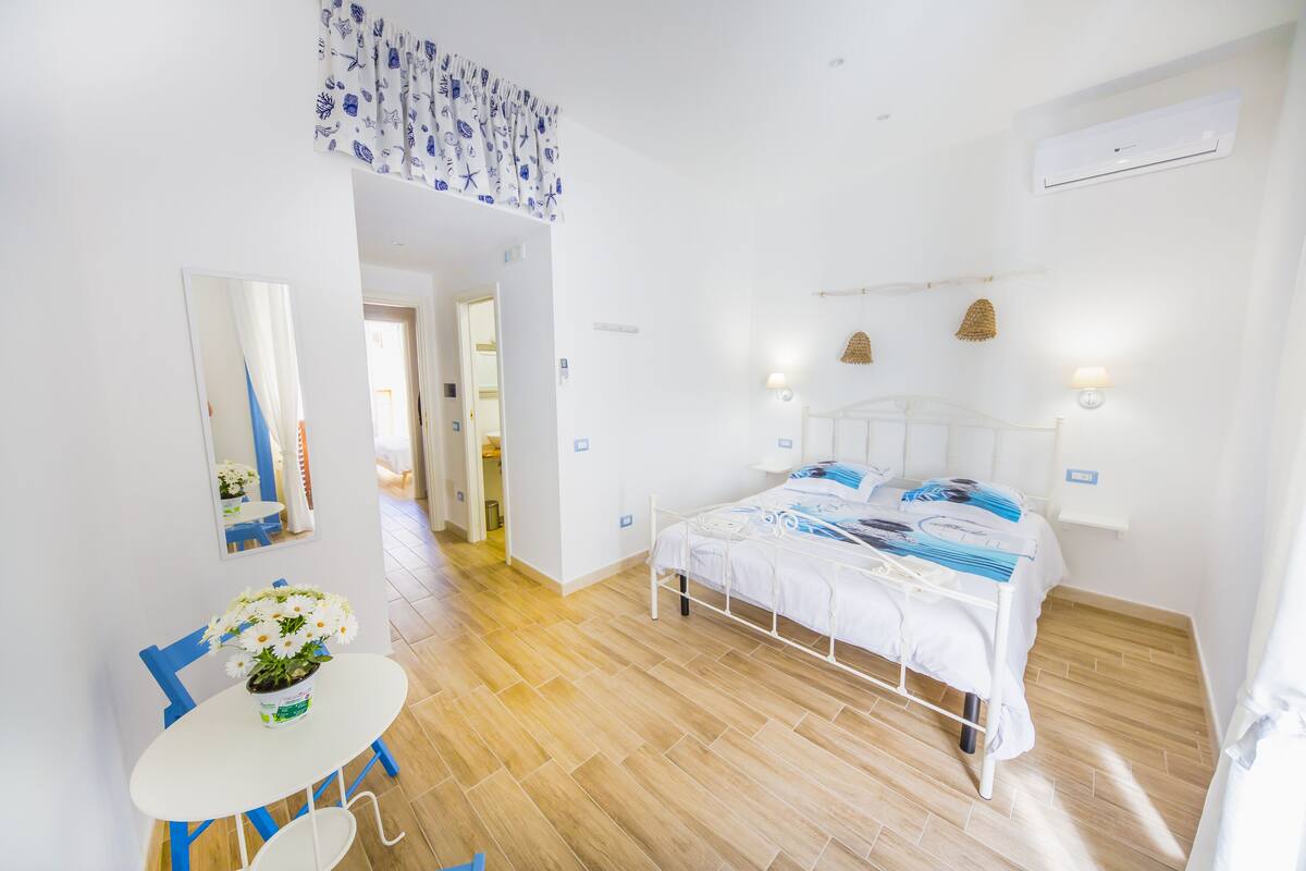 Una habitación luminosa y blanca con una cama, una mesita con una planta en maceta y un espejo en la pared.