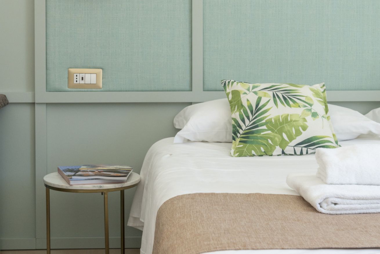 Een net opgemaakt bed met witte lakens, een sierkussen met bladpatroon en een opgevouwen handdoek ernaast. Een klein rond tafeltje met een boek en een afstandsbediening staat naast het bed.