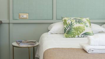 Een net opgemaakt bed met witte lakens, een sierkussen met bladpatroon en een opgevouwen handdoek ernaast. Een klein rond tafeltje met een boek en een afstandsbediening staat naast het bed.