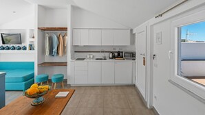 Une cuisine moderne avec des meubles blancs, un évier, une bouilloire et un four à micro-ondes. On y trouve une table à manger en bois sur laquelle est posé un saladier rempli de fruits.