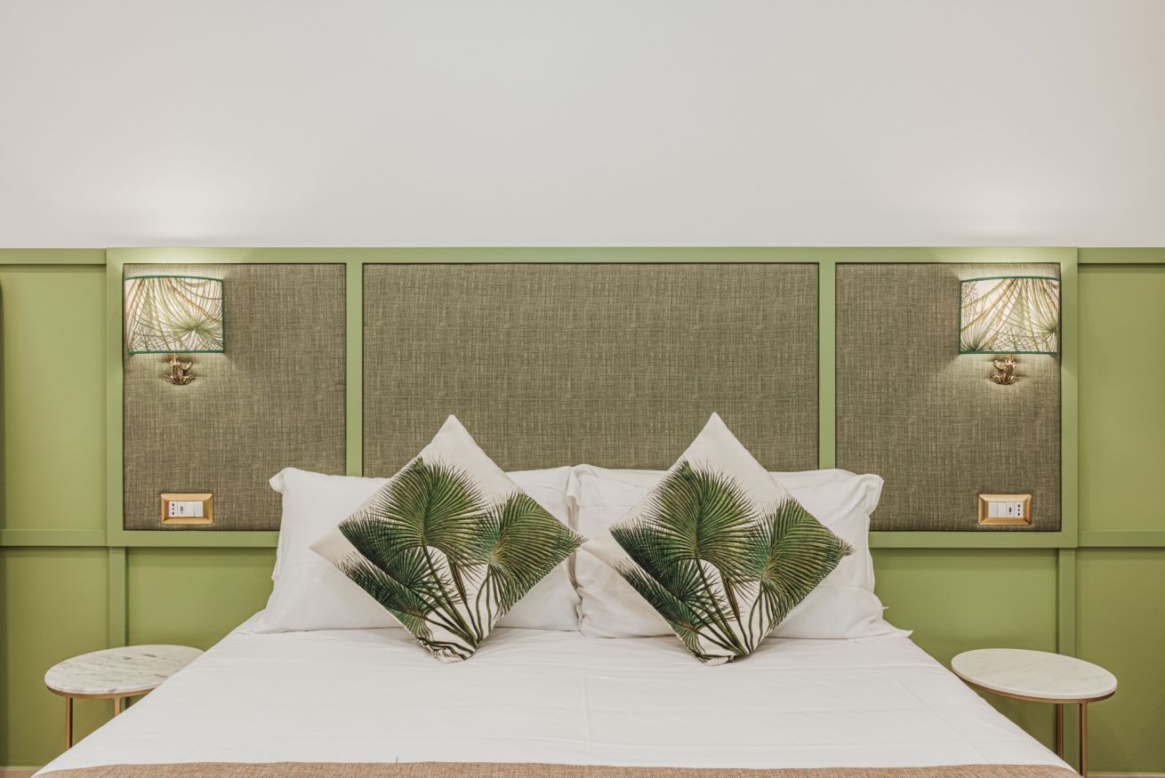 Una cama bien hecha con sábanas blancas y dos almohadas decorativas con diseños de palmeras. La habitación tiene paredes verdes y dos lámparas fijadas a la pared.