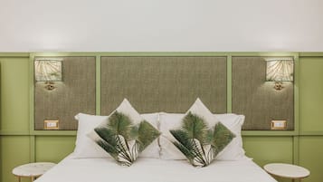 Una cama bien hecha con sábanas blancas y dos almohadas decorativas con diseños de palmeras. La habitación tiene paredes verdes y dos lámparas fijadas a la pared.