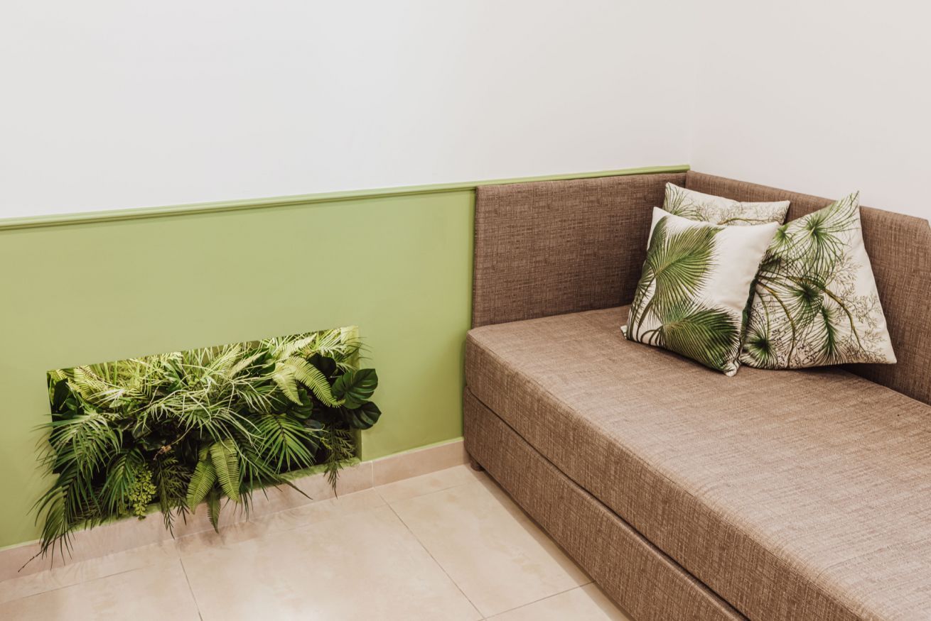 Rincón de una habitación con pared verde, un sofá marrón con cojines estampados y un arreglo de plantas colgado en la pared.