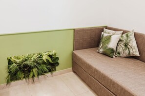 Rincón de una habitación con pared verde, un sofá marrón con cojines estampados y un arreglo de plantas colgado en la pared.
