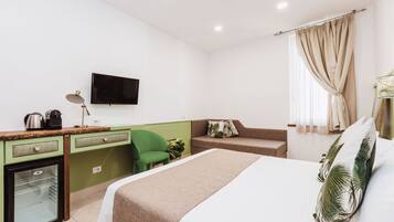 Une chambre d’hôtel avec un lit, un bureau avec un ordinateur, un petit réfrigérateur, une chaise verte, un canapé et un téléviseur fixé au mur.