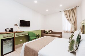 Habitación de hotel con cama, escritorio con computadora, refrigerador pequeño, una silla verde, sofá y televisor en la pared.