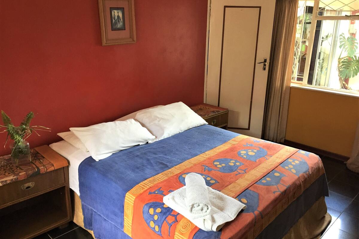 Ein Bett mit einer blauen und orangefarbenen Musterdecke, weißen Kissen und einem weißen Handtuch. Eine kleine Tisch mit einer Pflanze befindet sich links neben dem Bett. Rechts vom Bett ist eine Tür zu sehen, und ganz rechts ein Fenster mit Vorhängen.