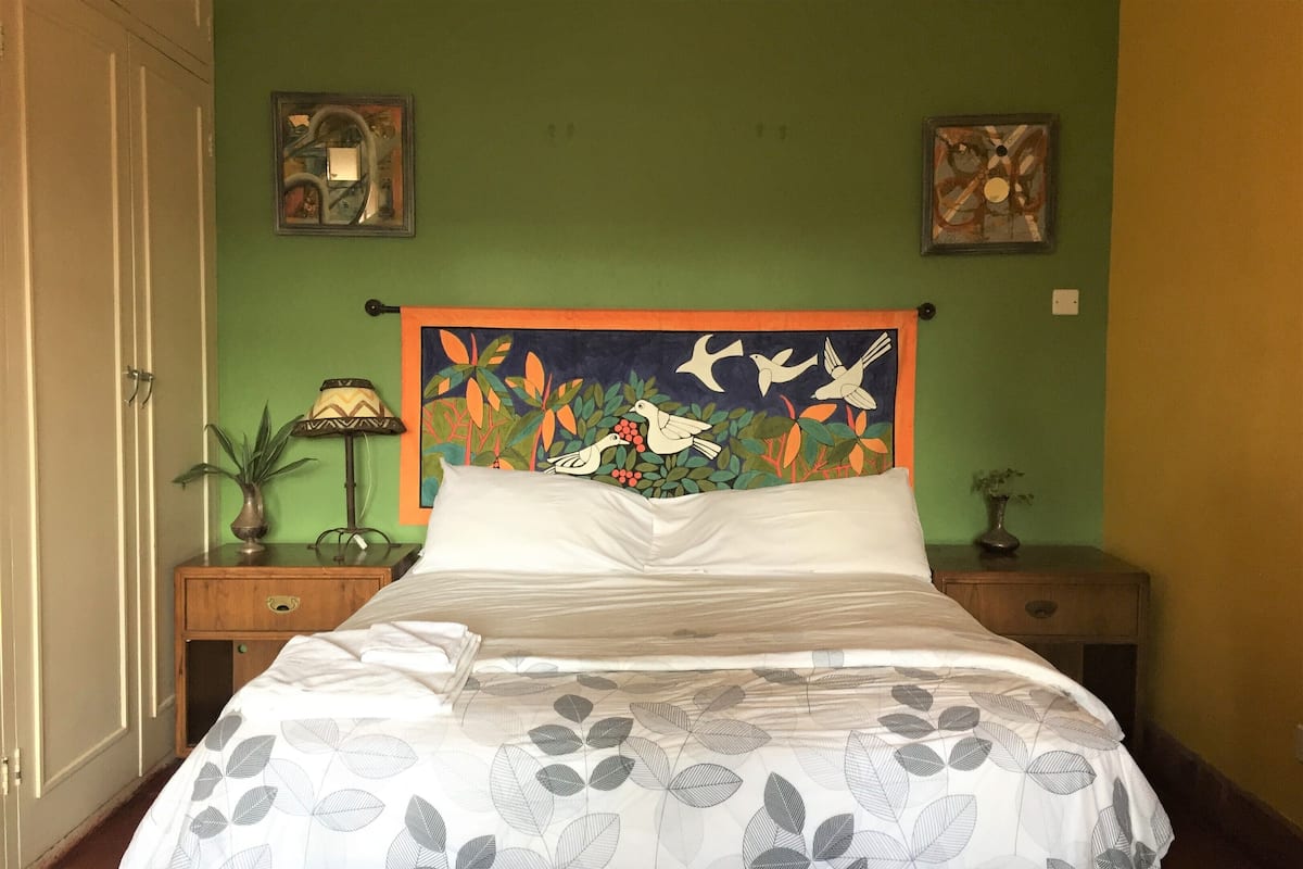 Ein Bett mit einem deckblatt mit laubmuster, ein kopfteil mit einem bild von vögeln und grünpflanzen, und zwei nachttische mit lampen und kleinen vasen.