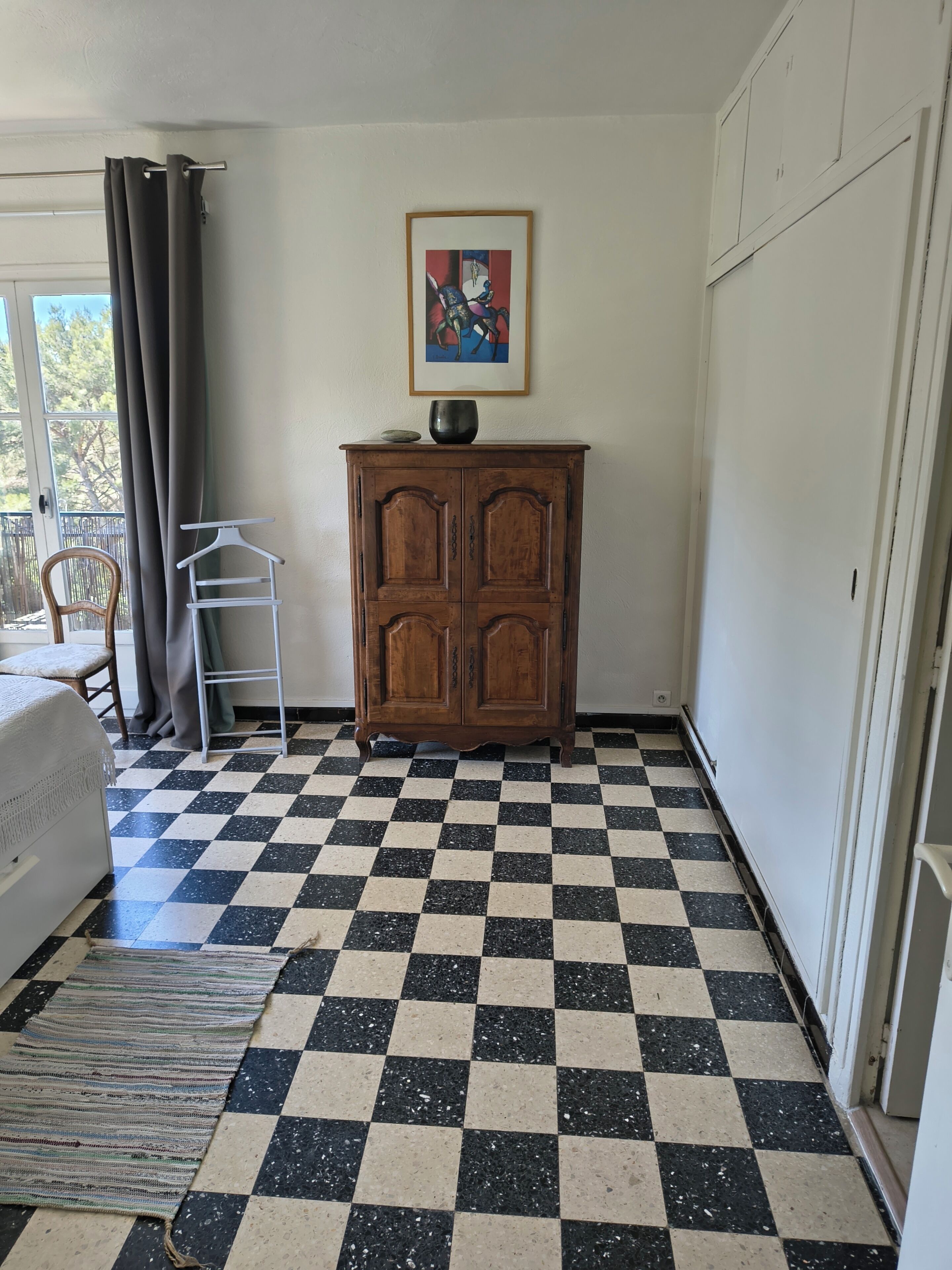 Ein Zimmer mit Schachbrettfußboden, einem Holzkoffer, einem gerahmten Bild und einem Fenster mit Vorhängen.