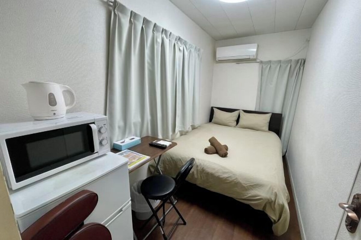 A small, clean room with a bed, a desk with a microwave, a chair, and a window with curtains.