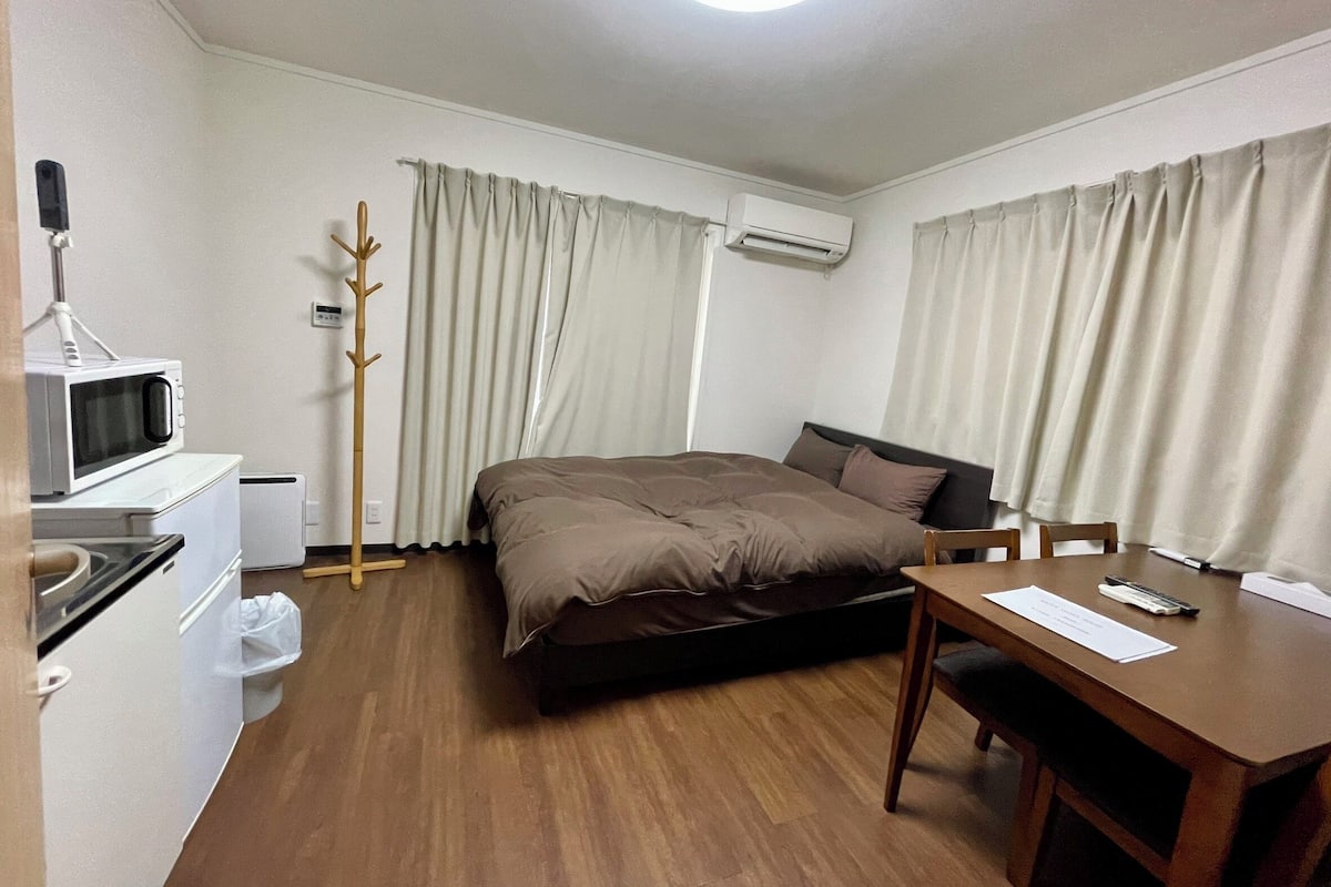 A hotel room with a bed, a desk, a chair, a microwave, a heater, and a coat rack.