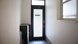 A narrow hallway with a black door featuring horizontal blinds, a visible emergency exit sign, and a window on the right. - Happy Osaka House (Osaka)