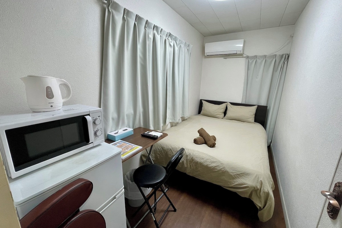 A compact hotel room with a bed, a desk with a kettle, a microwave, a chair, and an air conditioner.