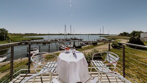 A table with a white tablecloth, two white chairs, a bowl of fruit, a bottle of wine, and a glass on a terrace overlooking a body of water with boats and a dock. - Apartment in the harbor (Puddemin)