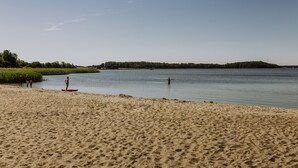 A sandy beach with a calm body of water, a small island in the distance, and a few people in the water. - Apartment in the harbor (Puddemin)
