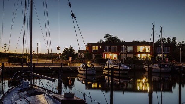 A marina at dusk with boats docked, a building with illuminated windows, and calm water reflecting the lights. - Apartment in the harbor (Puddemin)