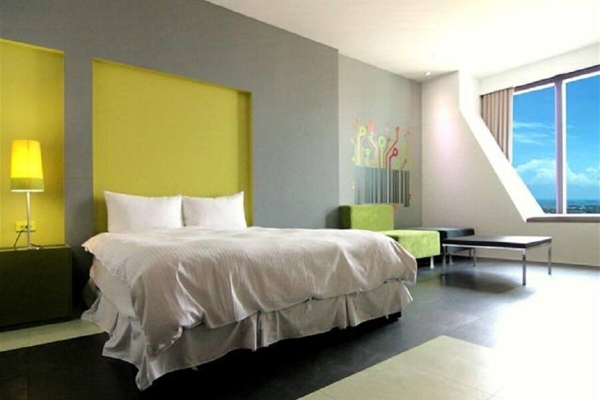 A modern hotel room with a large bed, a desk, and a view of the city through a large window.