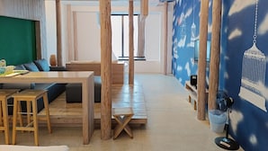 Una habitación con piso de madera, pared verde, una mesa de madera con taburetes y pared azul con un mural de un pájaro.