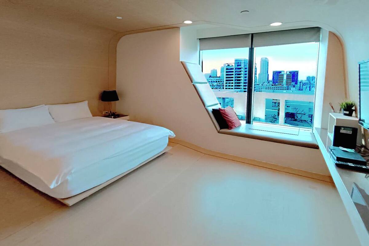 A modern bedroom with a bed, a nightstand, a desk, and a large window offering a city view.