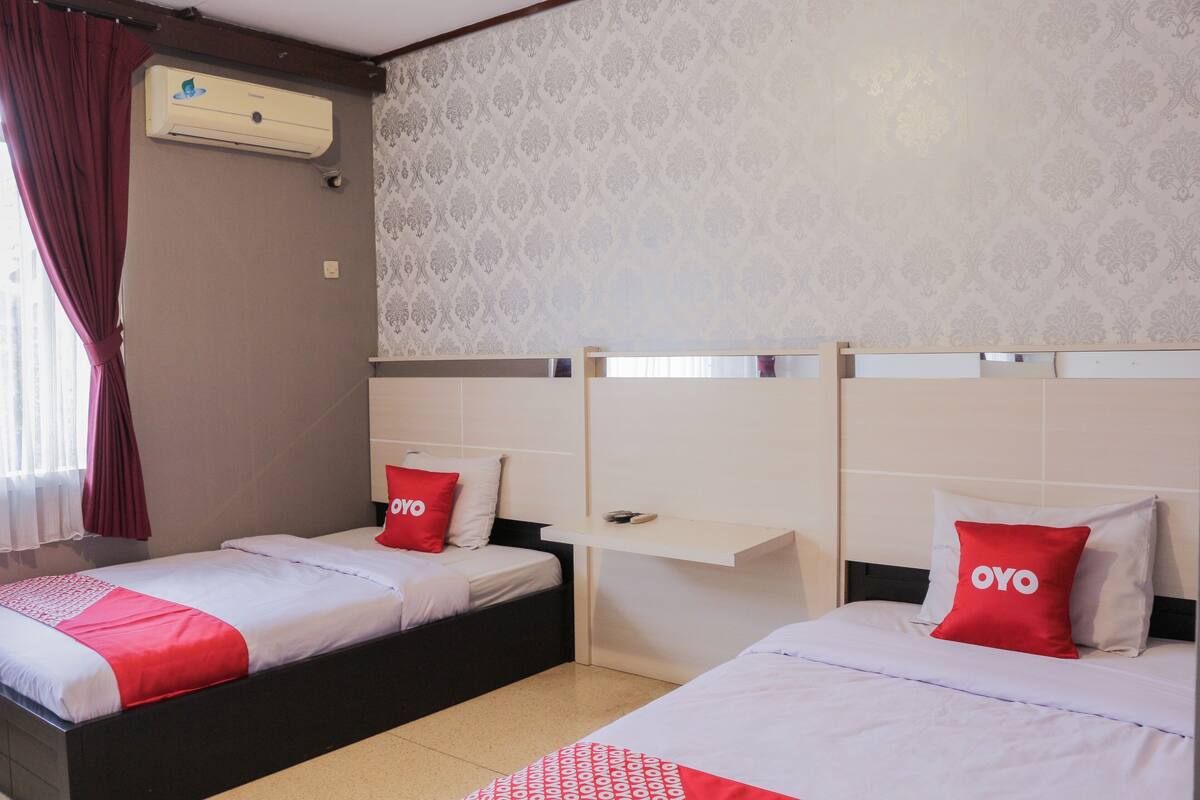 Habitación de hotel con dos camas, cada una con un cojín rojo de OYO, un cabecero blanco y una pequeña estantería.