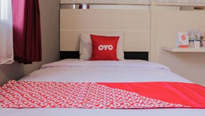 A hotel room with a bed, a red and white patterned bedspread, a red pillow with "OYO" text, a red curtain, and a shelf with toiletries.