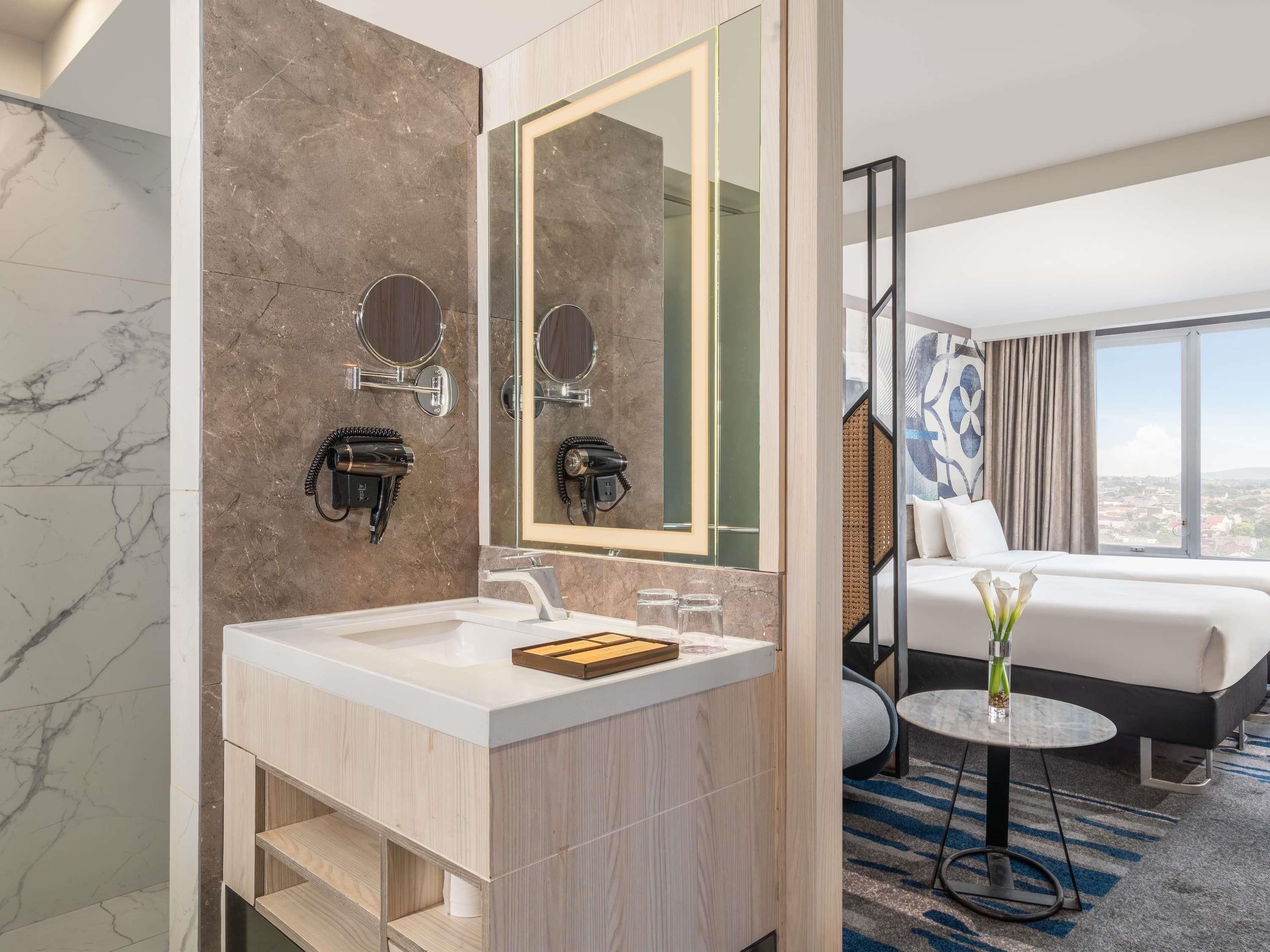 A modern hotel bathroom with a marble sink, a large mirror, and a hairdryer mounted on the wall.