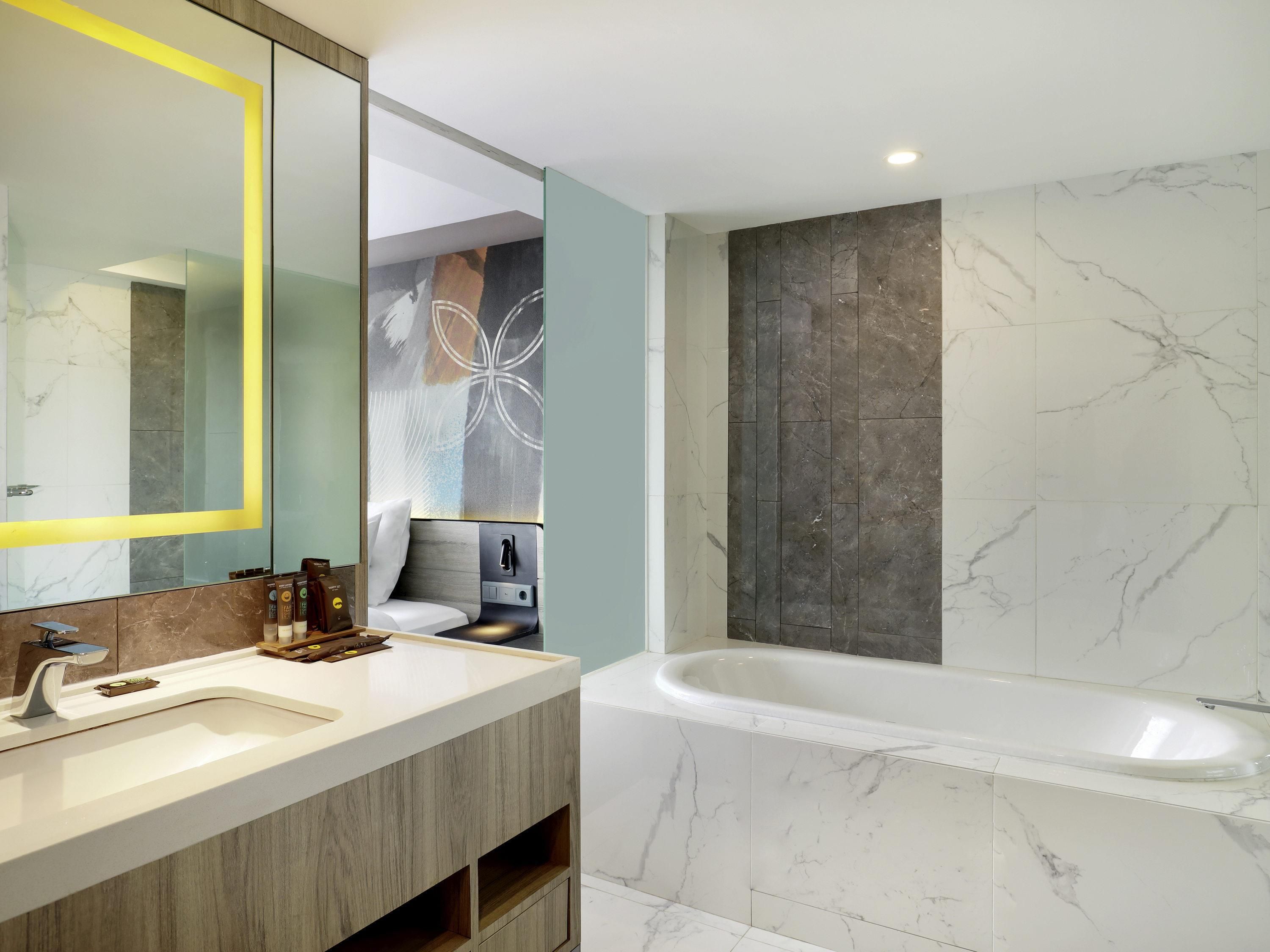A modern bathroom with a large mirror, a white sink, a wooden vanity, a bathtub, and marble tiles.