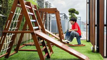 A child playing on a wooden slide structure in a garden area with artificial grass and a stone wall in the background.
