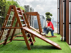 Een kind speelt op een houten glijbaanconstructie in een tuin met kunstgras en een stenen muur op de achtergrond.