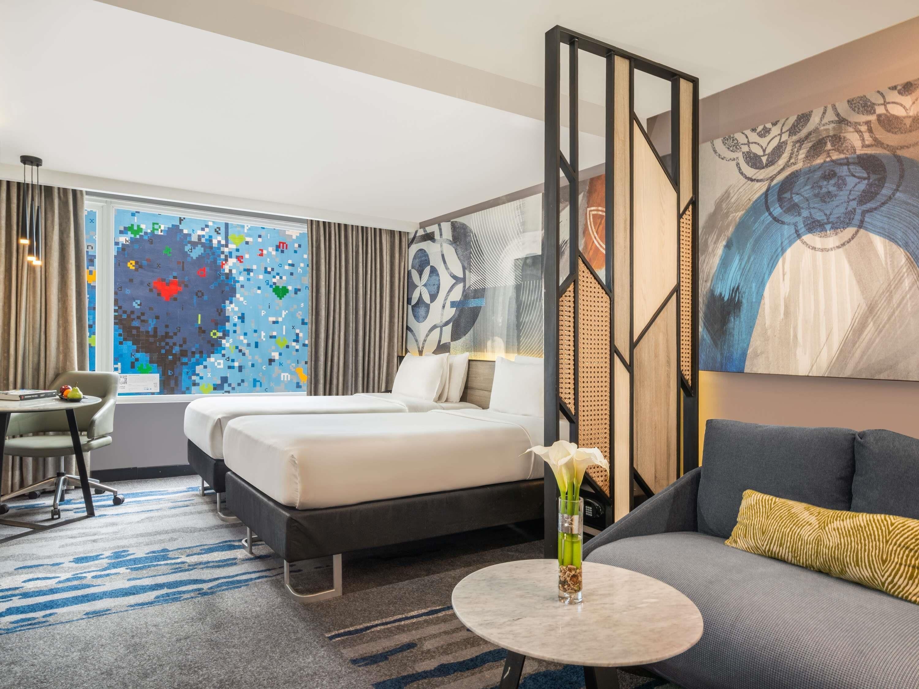 A hotel room with a bed, a sofa, a desk, and a large window with a colorful abstract wall art.