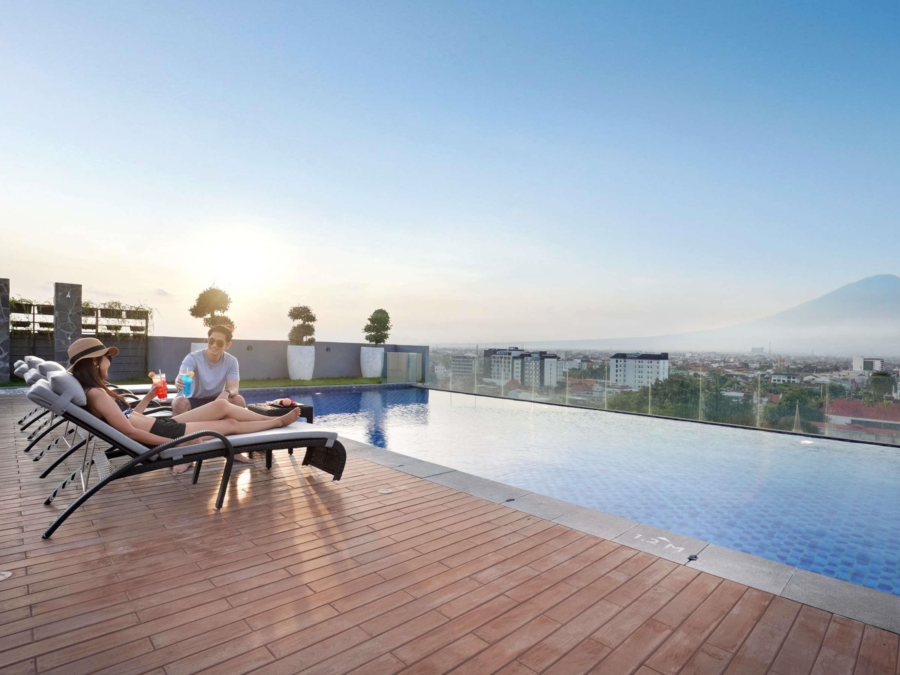 Una piscina infinita en la azotea con sillones de descanso, una terraza de madera y vistas a la ciudad.