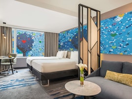 A hotel room with a bed, a desk, a chair, a sofa, and a large window with curtains.