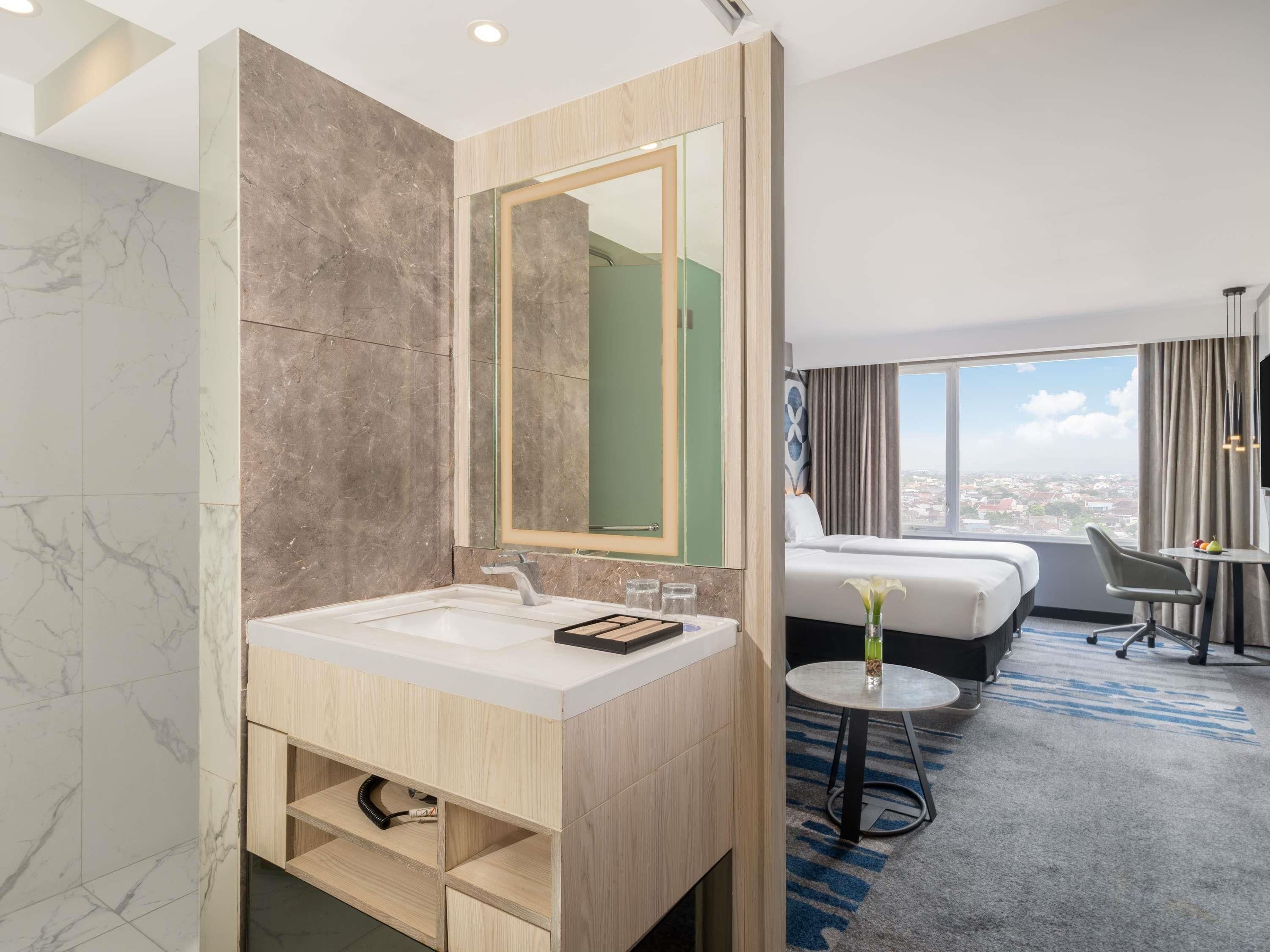 A hotel bathroom with a marble vanity, a large mirror, a bed with a view, and a small table with a vase.