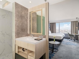 A hotel bathroom with a marble vanity, a large mirror, a bed with a view, and a small table with a vase.