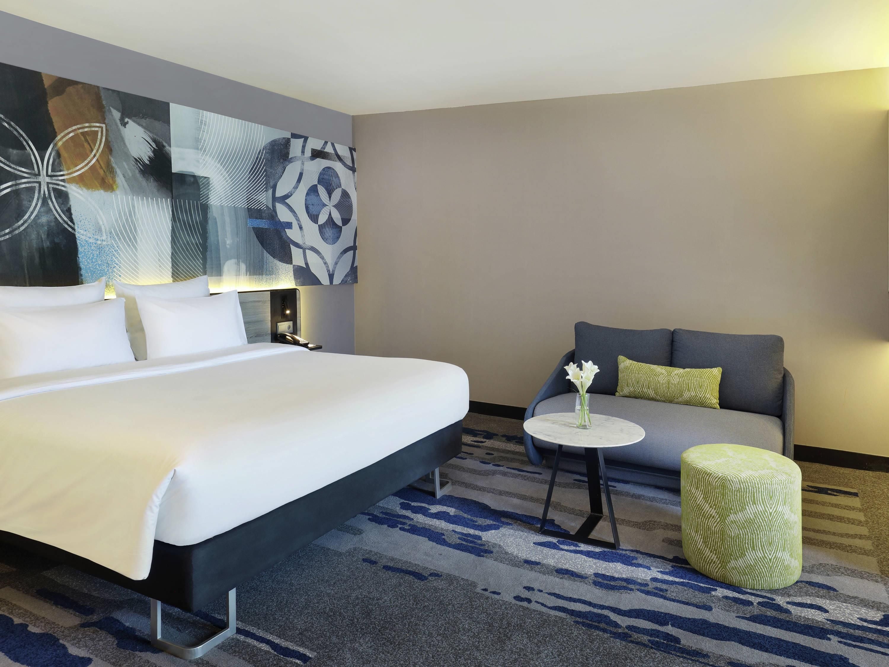 Een hotelkamer met een groot bed, een slaapbank, een klein rond tafeltje met een vaas en een kamerbreed tapijt met dessin.