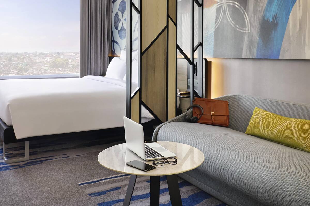 Ein Hotelzimmer mit einem Bett, einer Couch, einem runden Tisch mit einem Laptop und Gläsern sowie Blick auf die Stadt.