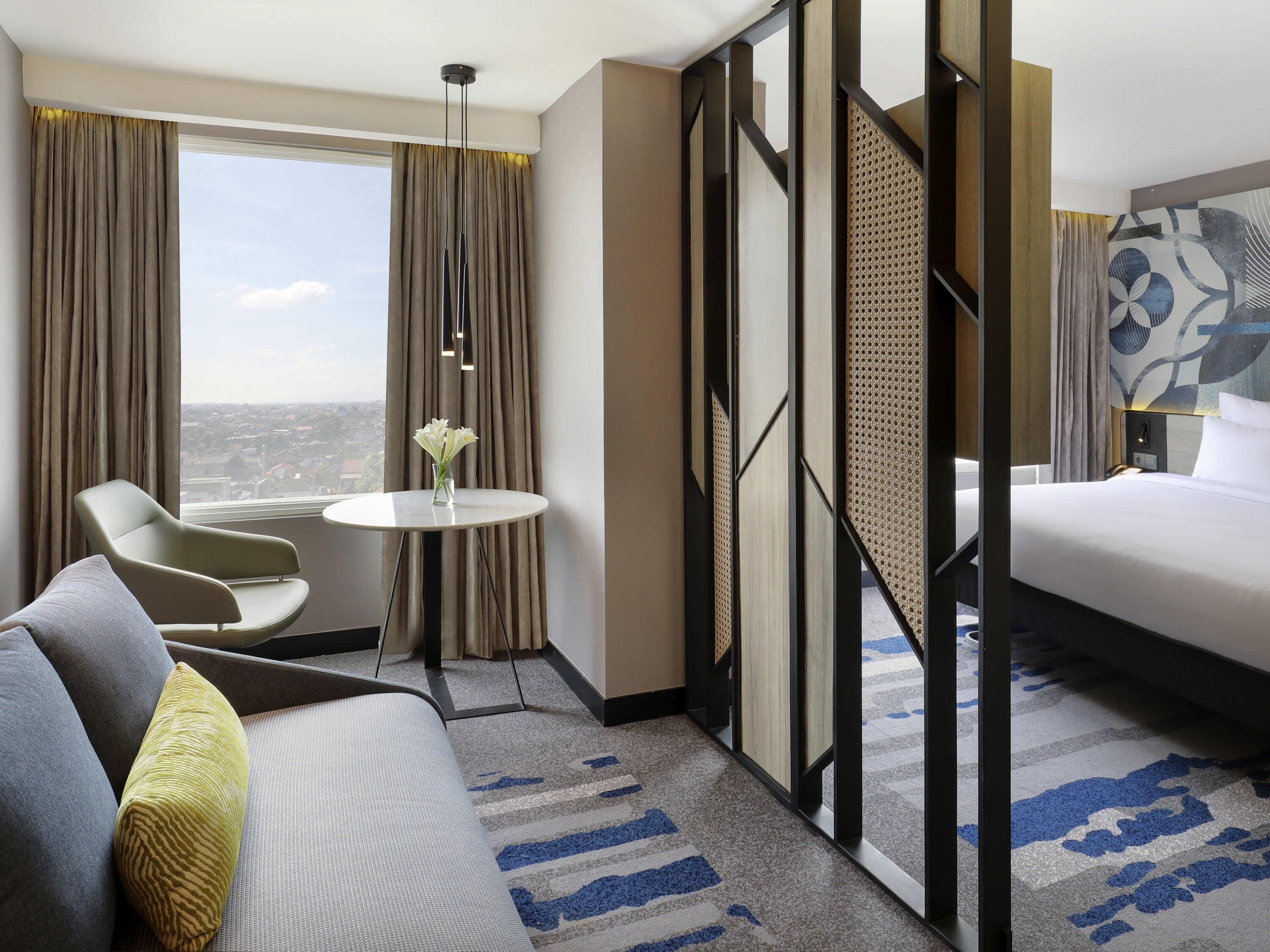 A hotel room with a bed, a sofa, a chair, a small table, and a view of the city.