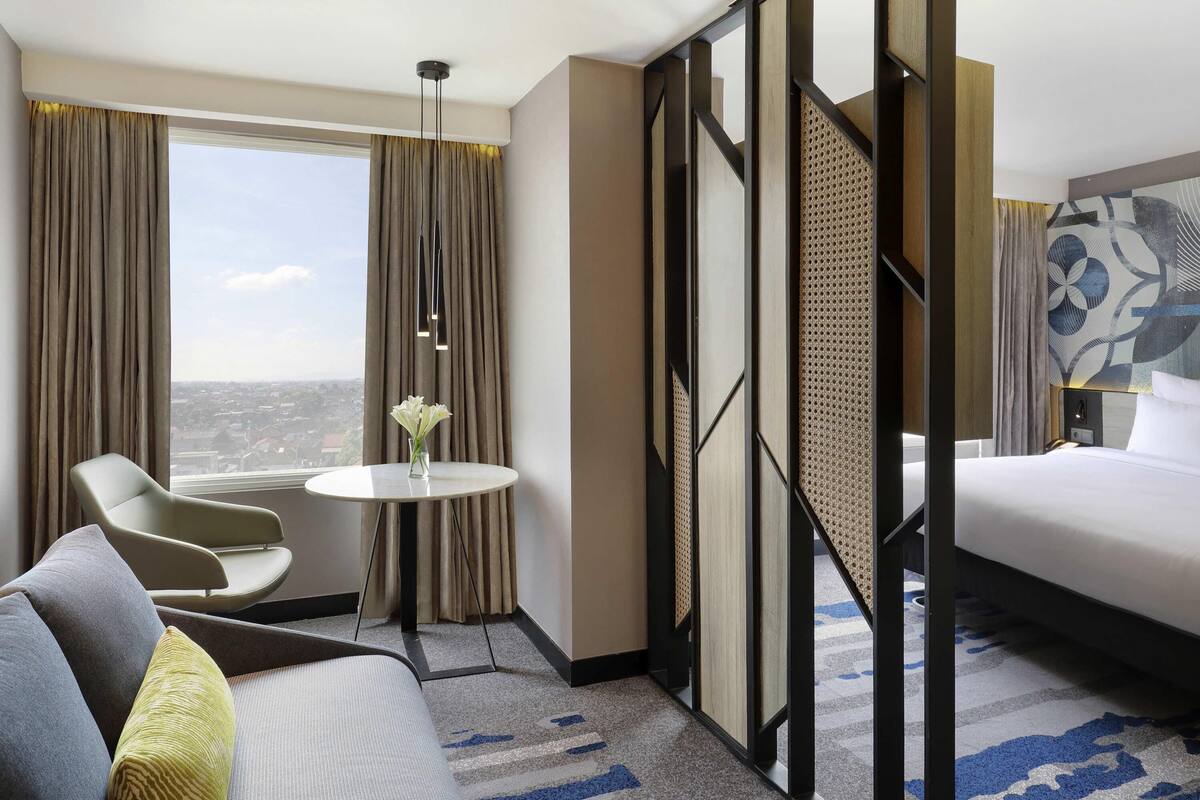 A hotel room with a bed, a sofa, a chair, a small table, and a view of the city.