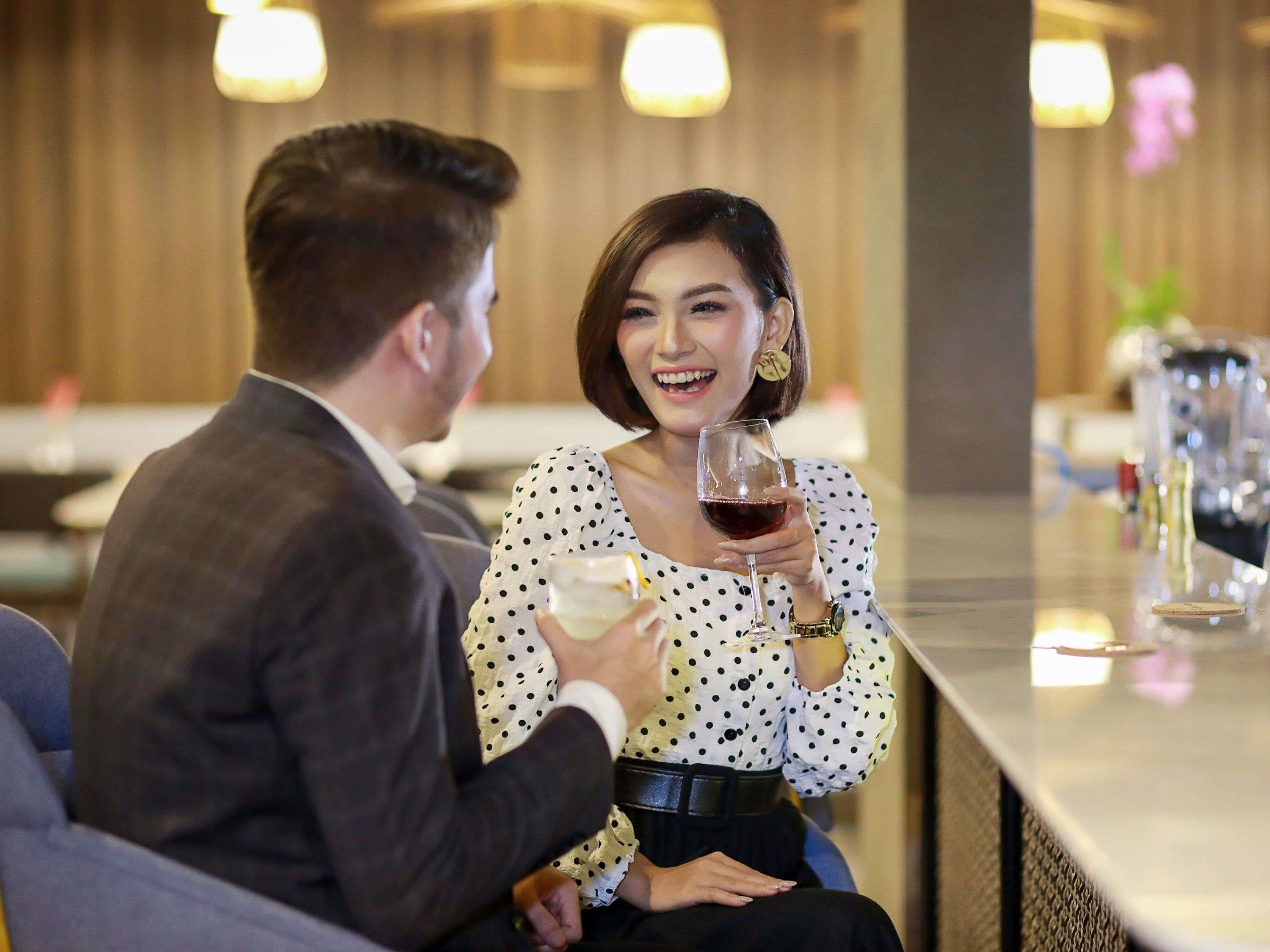 Una pareja sentada en un bar, cada uno con una bebida en la mano, un reloj sobre la mesa y una botella de vino.