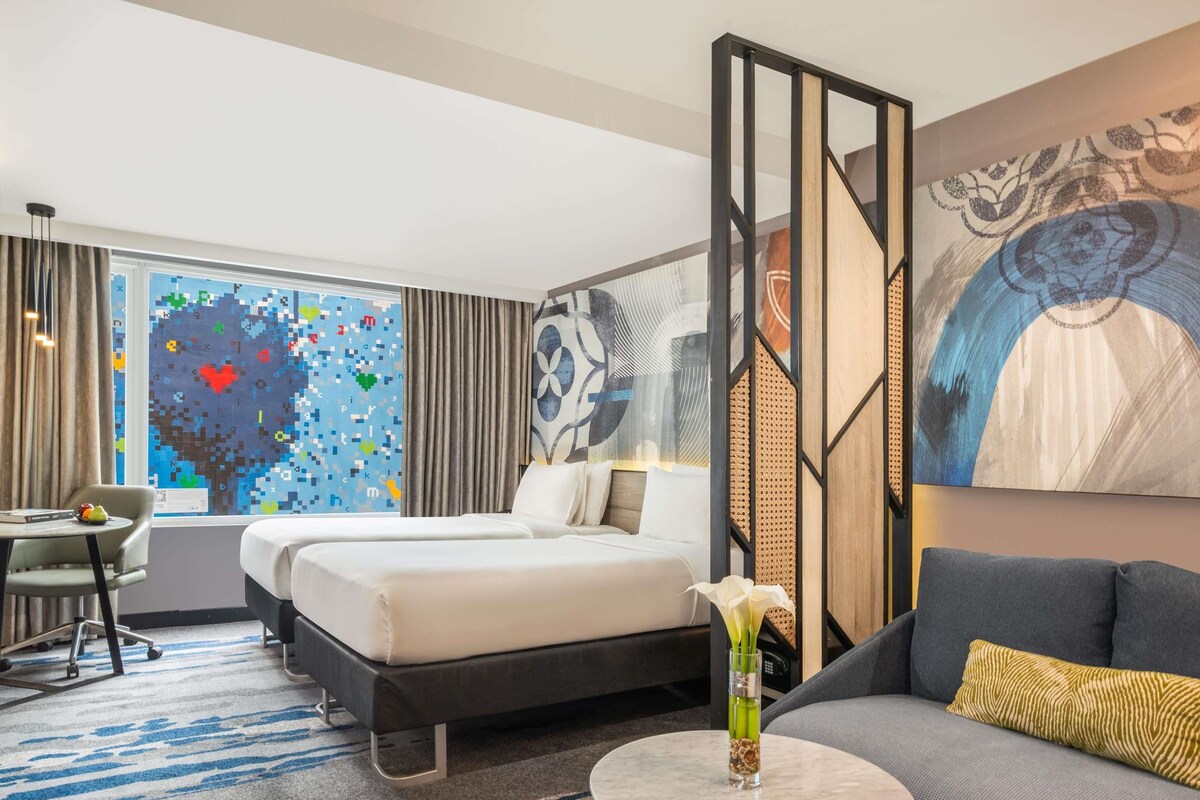 A hotel room with a bed, a sofa, a desk, and a large window with a colorful abstract wall art.