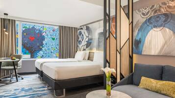 A hotel room with a bed, a sofa, a desk, and a large window with a colorful abstract wall art.